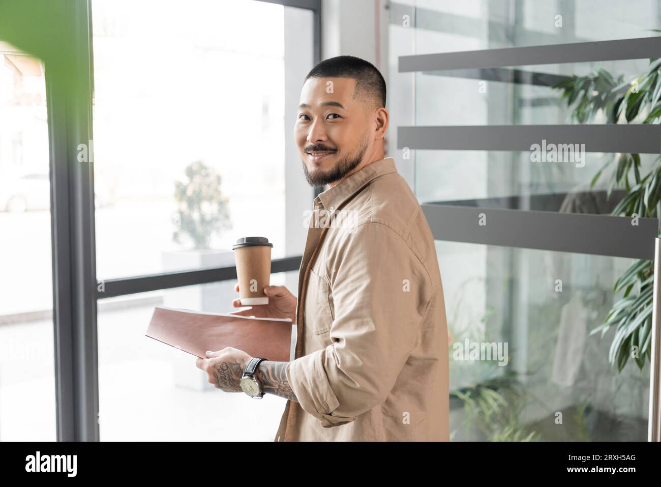homme d'affaires asiatique heureux avec tatouage souriant et marchant dans le bureau avec dossier et tasse de papier Banque D'Images