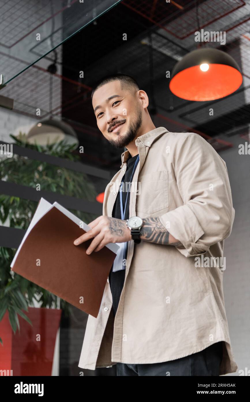 joyeux travailleur de bureau asiatique avec tatouage tenant le dossier et regardant la caméra près de la porte vitrée Banque D'Images