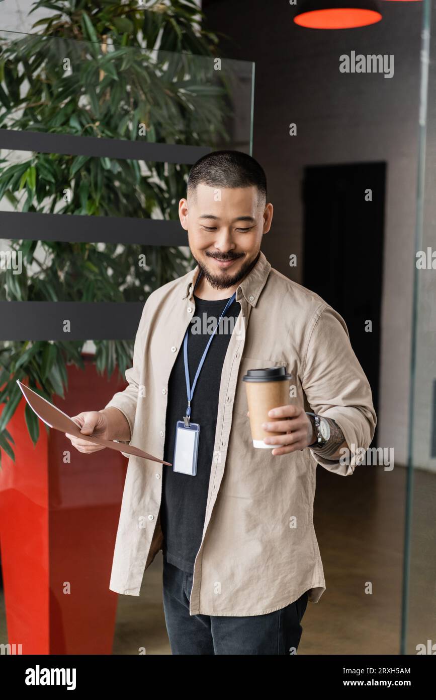 joyeux homme d'affaires asiatique avec tatouage souriant et entrant dans le bureau avec dossier et tasse de papier Banque D'Images