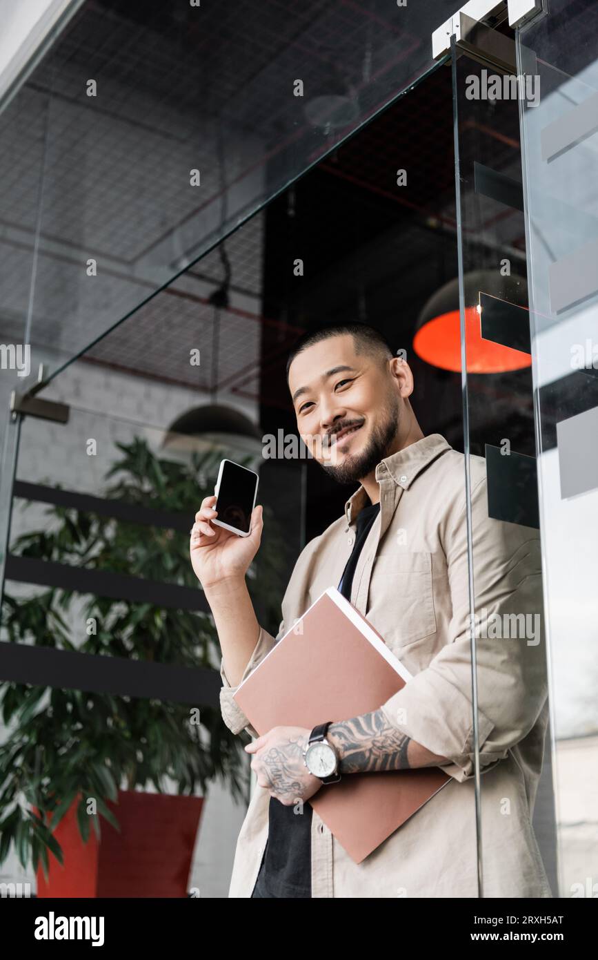 heureux homme d'affaires asiatique avec tatouage tenant le dossier et smartphone près de la porte en verre du bureau Banque D'Images