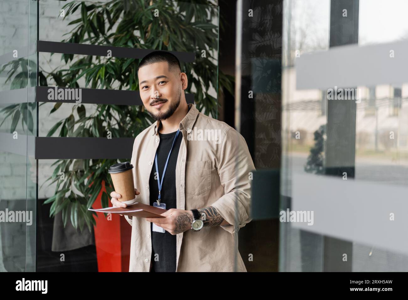 heureux homme d'affaires asiatique avec tatouage souriant et debout avec dossier et café dans la tasse de papier Banque D'Images