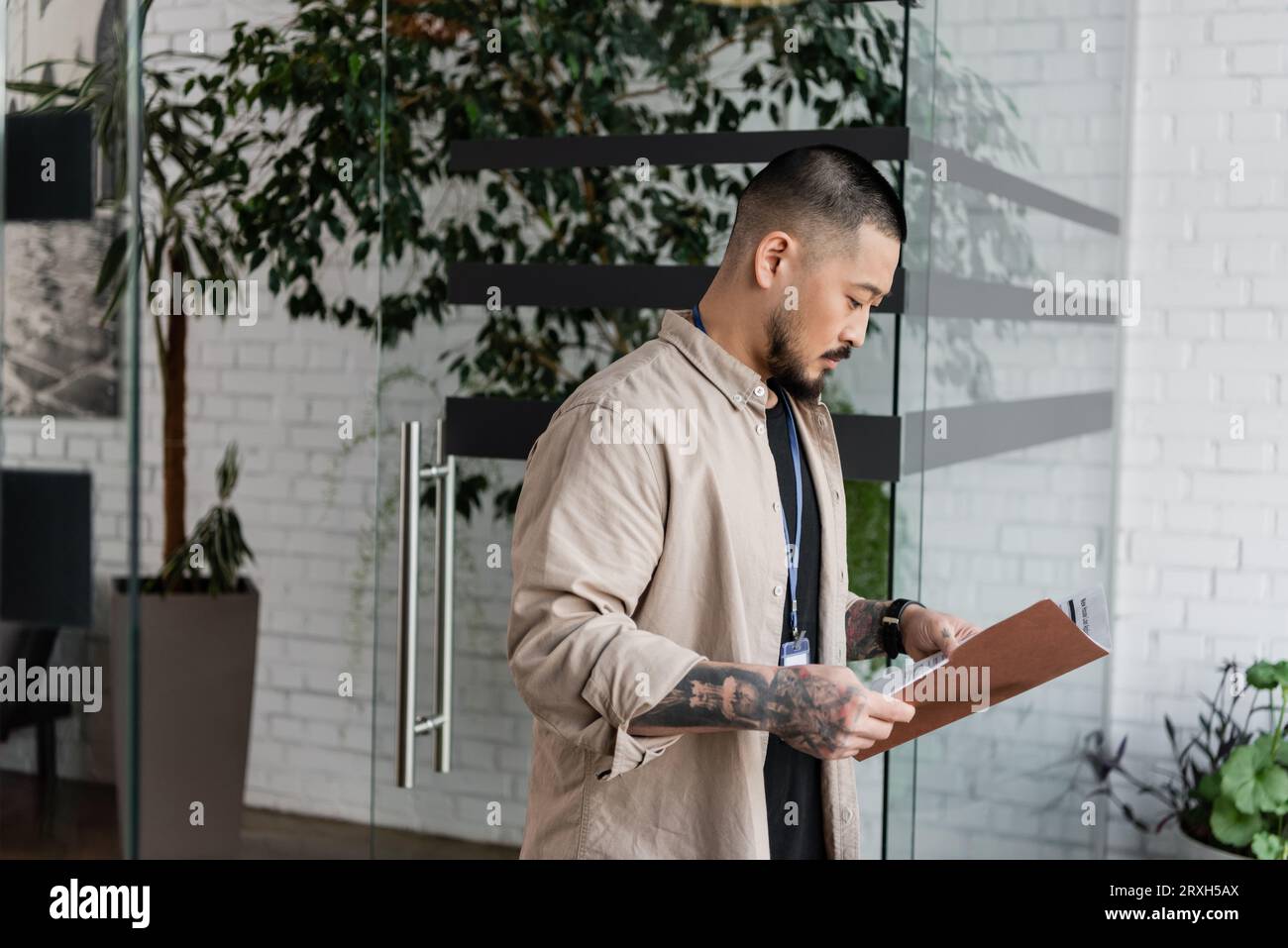 homme d'affaires asiatique concentré avec tatouage regardant le plan de démarrage à l'intérieur du dossier près de la porte en verre Banque D'Images