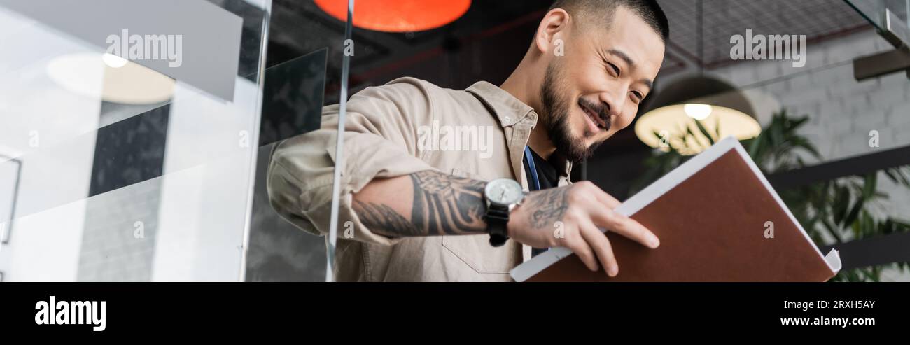 heureux homme d'affaires asiatique avec tatouage regardant les documents à l'intérieur du dossier près de la porte en verre, bannière Banque D'Images