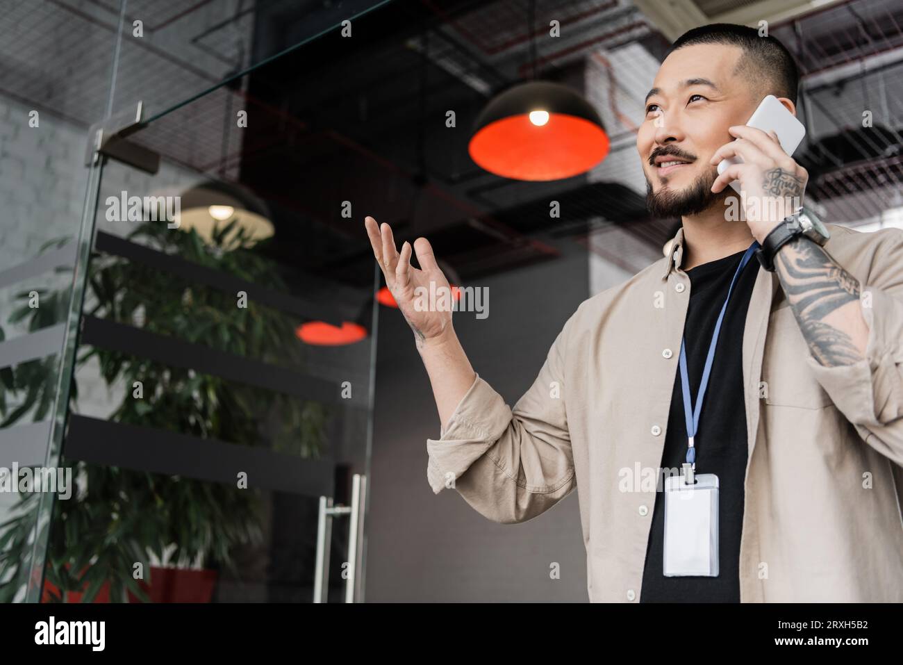 heureux homme d'affaires asiatique avec tatouage gesturant pendant l'appel téléphonique smartphone dans le bureau moderne Banque D'Images