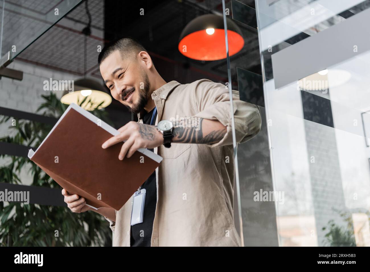 homme d'affaires asiatique heureux avec tatouage regardant les documents à l'intérieur du dossier près de la porte en verre, angle bas Banque D'Images
