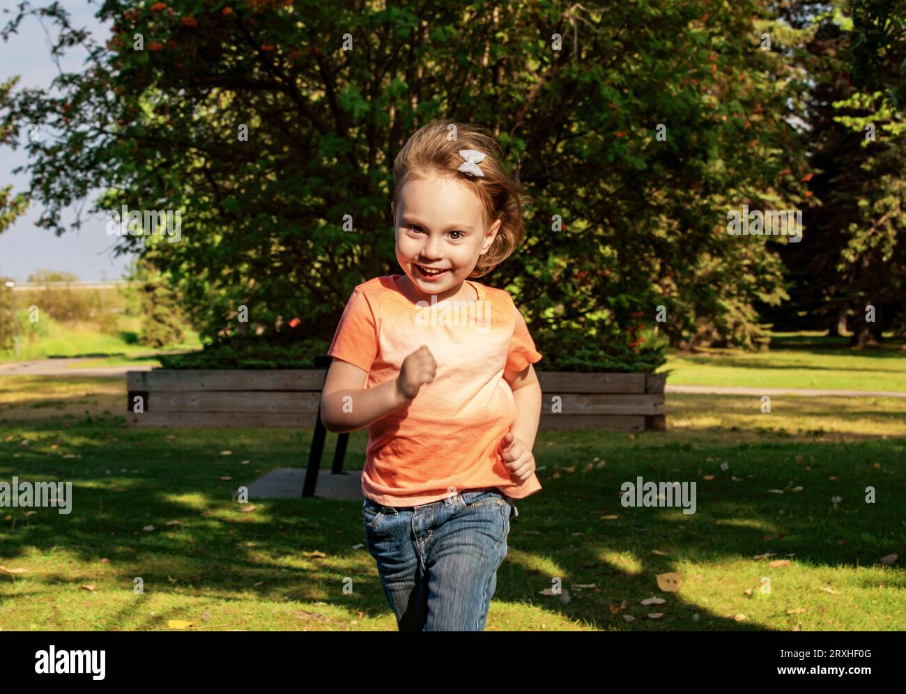 Fille d'enfant d'âge préscolaire courant vers la caméra dans la lumière du soleil avec un grand sourire sur son visage, dans un parc de la ville par un après-midi chaud d'automne Banque D'Images