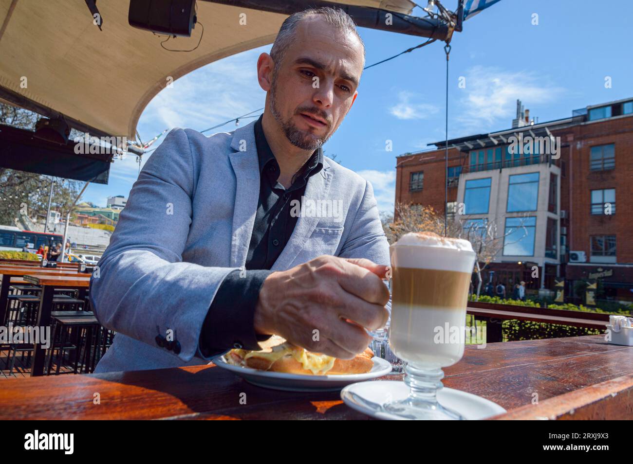 Homme d'affaires caucasien adulte dans des vêtements élégants, prenant une tasse de café cappuccino du comptoir, assis prenant le petit déjeuner à l'extérieur du restaurant. Banque D'Images