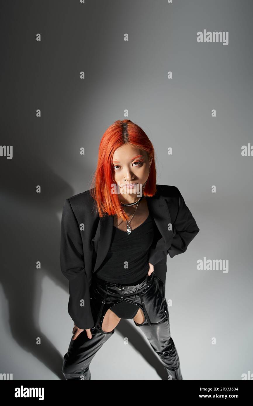 modèle asiatique élégant avec les cheveux rouges et pose piercing posant avec la main sur la hanche, debout dans le blazer Banque D'Images