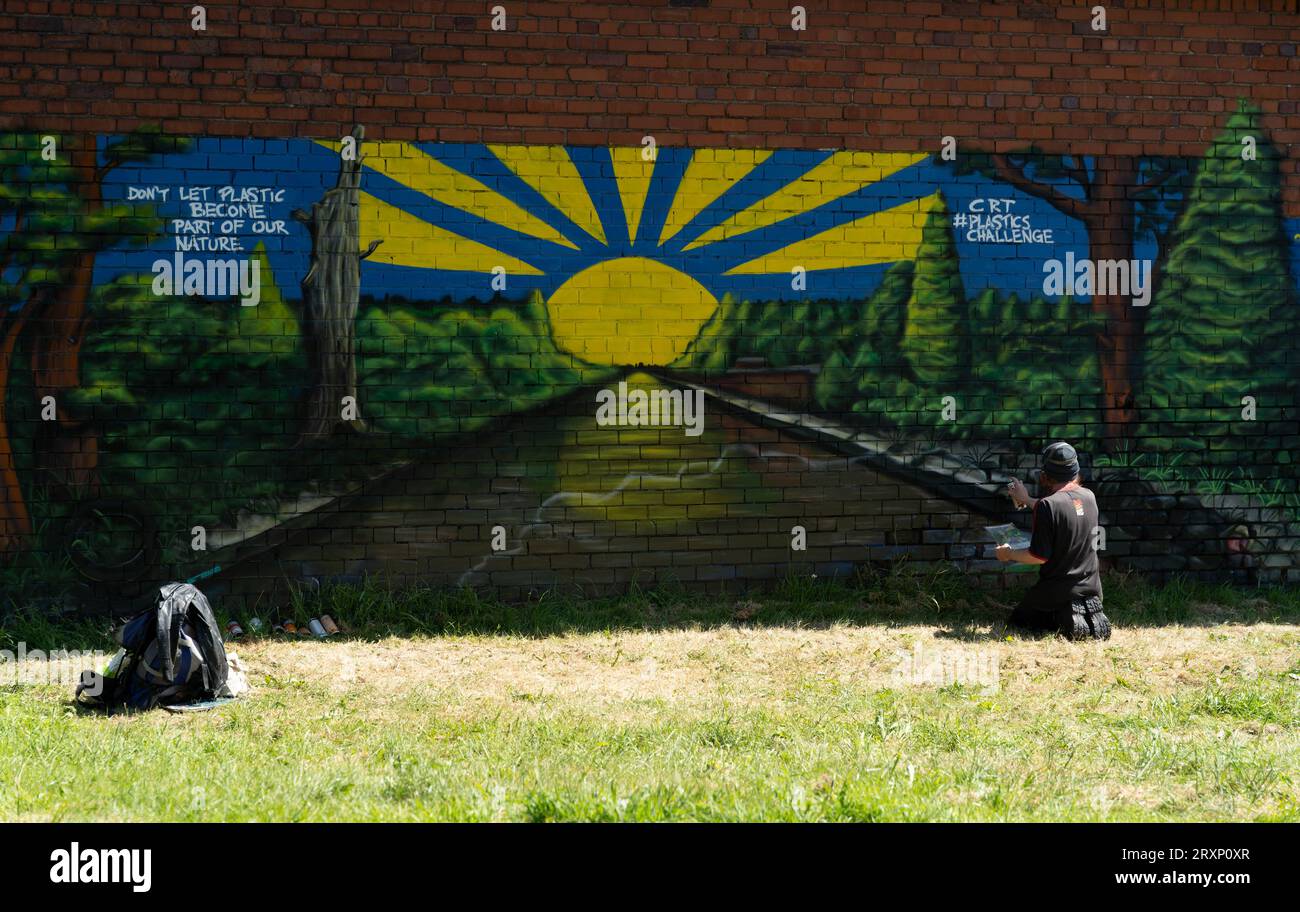 Artiste créant une murale environnementale sur un mur de briques avec un message anti-plastique et une scène de paysage au lever du soleil Banque D'Images