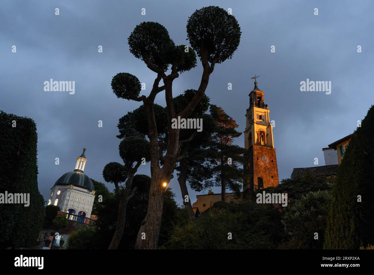 Clocher illuminé et bâtiment en forme de dôme au crépuscule dans le village de Portmeirion avec des arbres topiaires Banque D'Images Clocher illuminé et bâtiment en forme de dôme au crépuscule dans le village de Portmeirion avec des arbres topiaires Banque D'Images