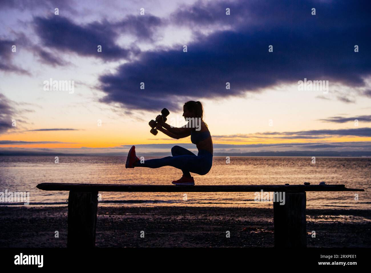 Silhouette de femme s'exerçant avec des haltères sur le dessus du banc côtier au crépuscule nuageux Banque D'Images