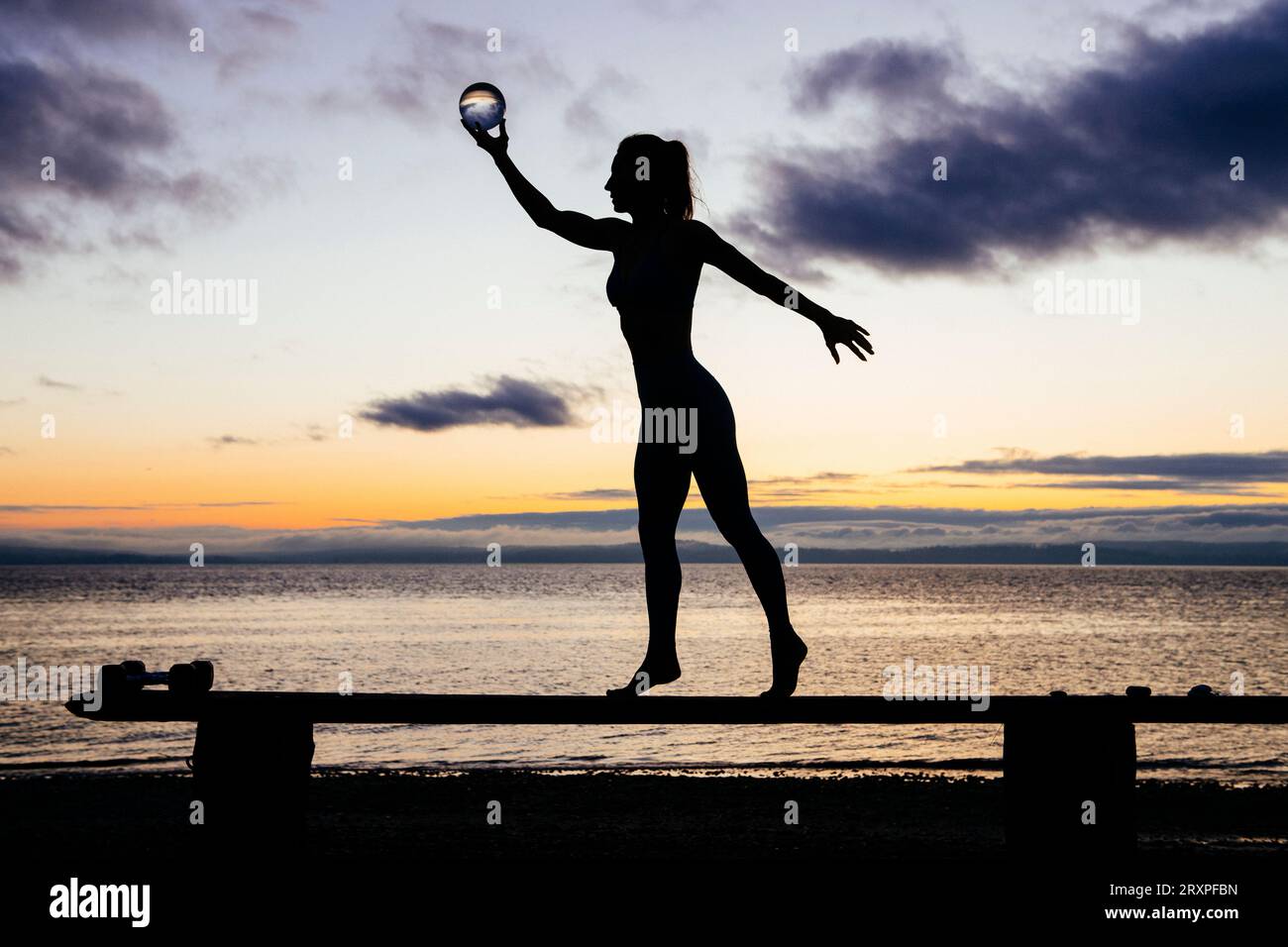 Silhouette de femme s'exerçant avec des haltères sur le dessus du banc côtier au crépuscule nuageux Banque D'Images