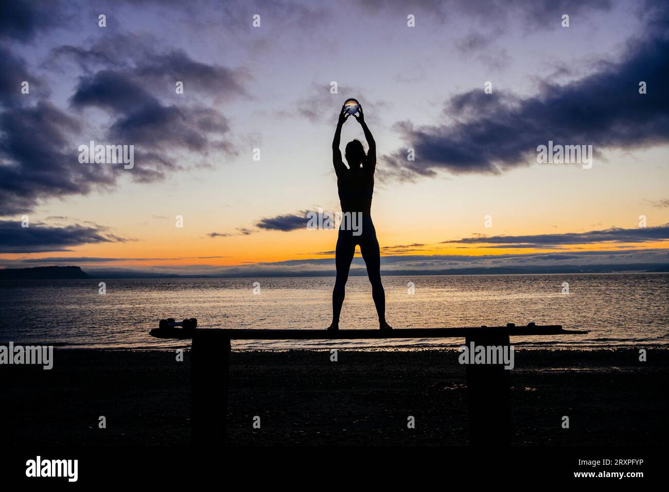 Silhouette de femme s'exerçant avec des haltères sur le dessus du banc côtier au crépuscule nuageux Banque D'Images