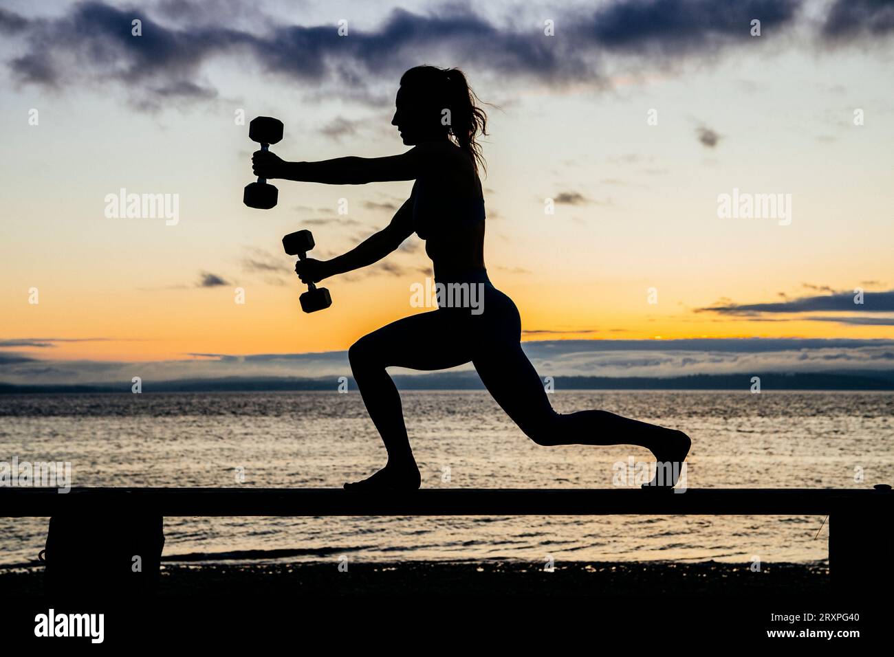 Silhouette de femme s'exerçant avec des haltères sur le dessus du banc côtier au crépuscule nuageux Banque D'Images