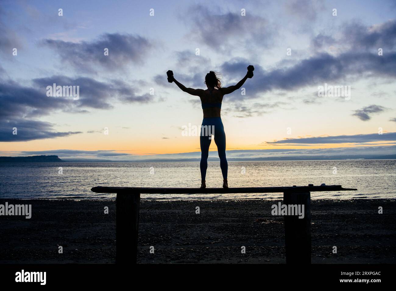 Silhouette de femme s'exerçant avec des haltères sur le dessus du banc côtier au crépuscule nuageux Banque D'Images