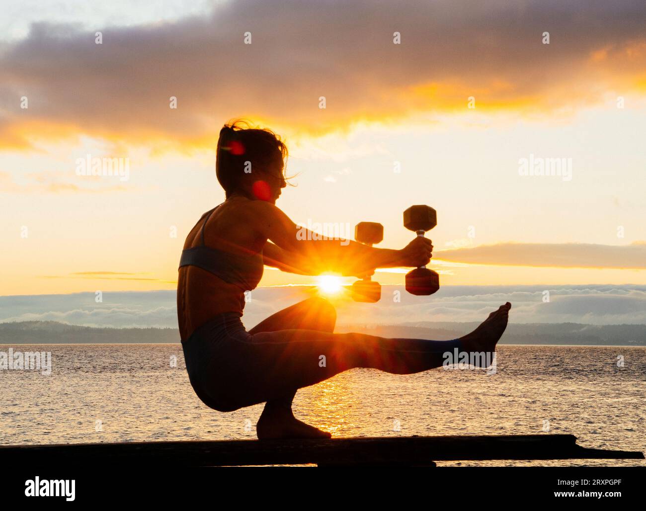 Silhouette de femme s'exerçant avec des haltères sur le dessus du banc côtier au crépuscule nuageux Banque D'Images