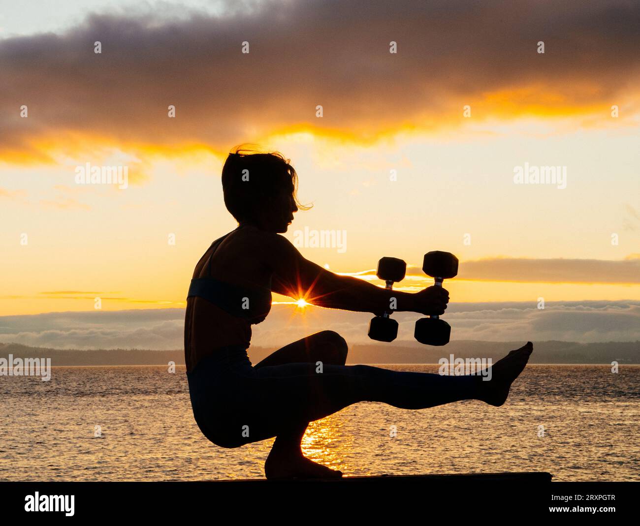 Silhouette de femme s'exerçant avec des haltères sur le dessus du banc côtier au crépuscule nuageux Banque D'Images