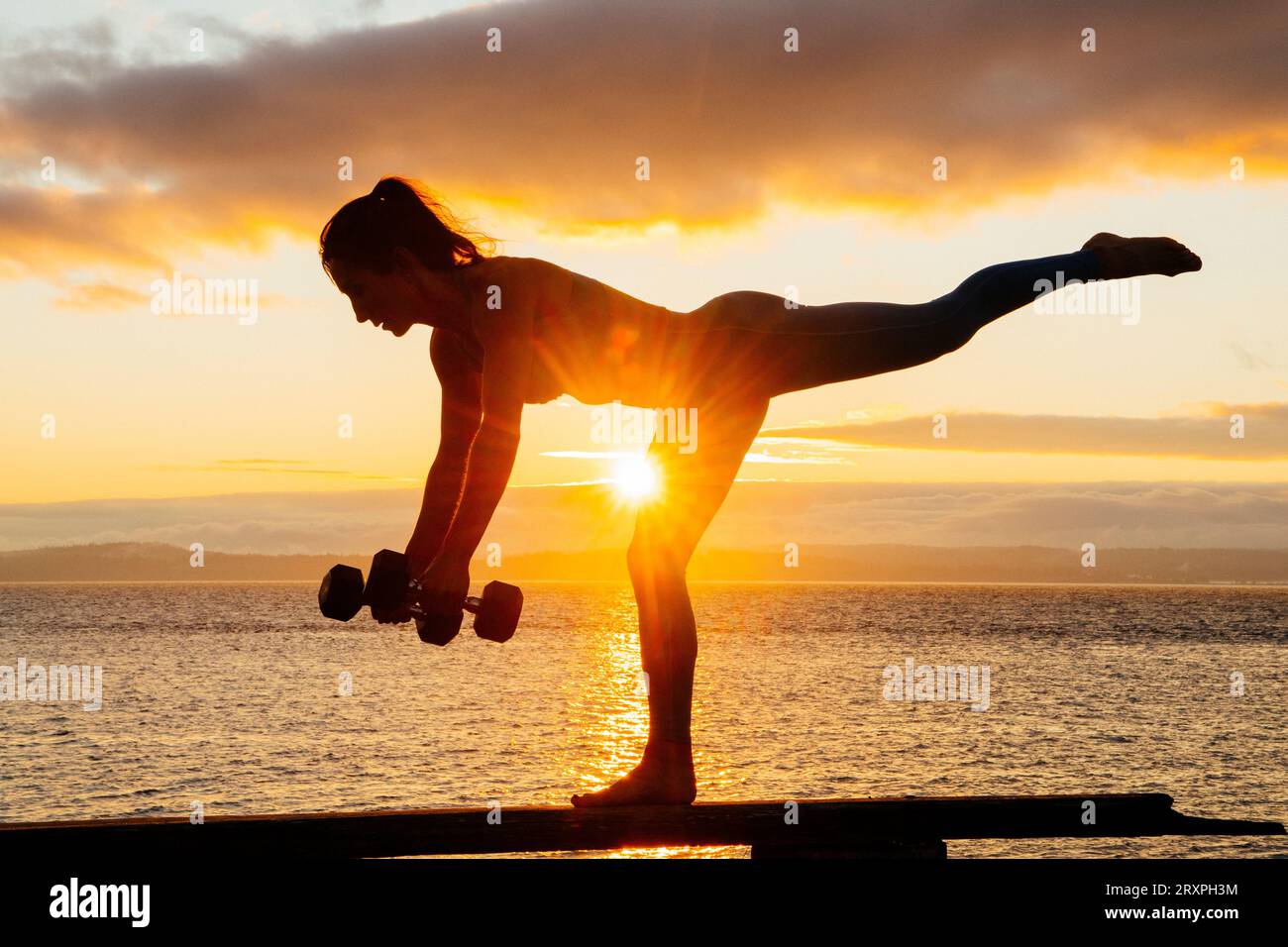 Silhouette de femme s'exerçant avec des haltères sur le dessus du banc côtier au crépuscule nuageux Banque D'Images