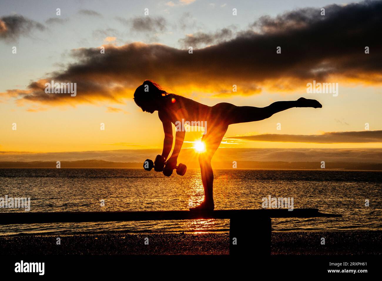 Silhouette de femme s'exerçant avec des haltères sur le dessus du banc côtier au crépuscule nuageux Banque D'Images