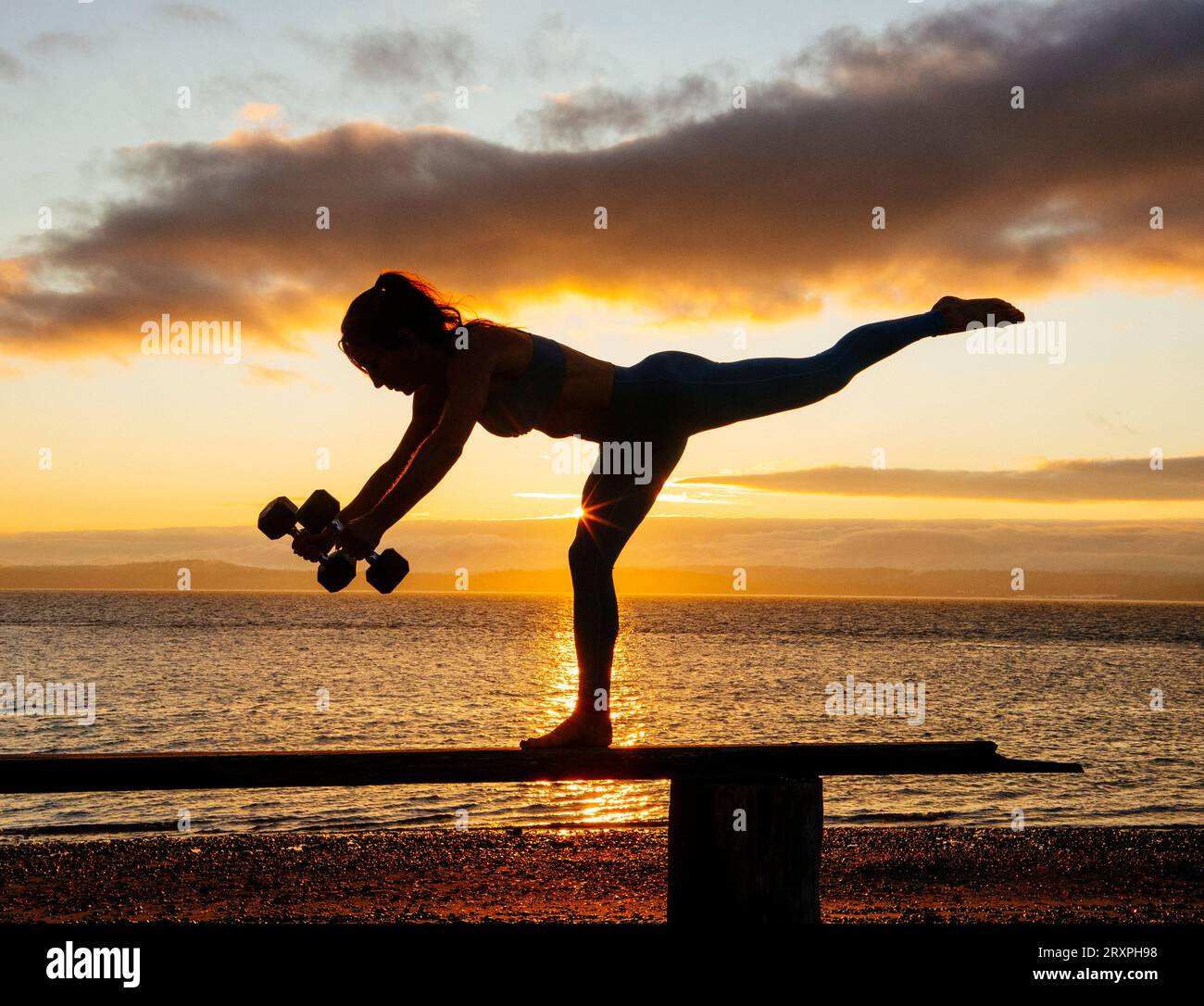 Silhouette de femme s'exerçant avec des haltères sur le dessus du banc côtier au crépuscule nuageux Banque D'Images