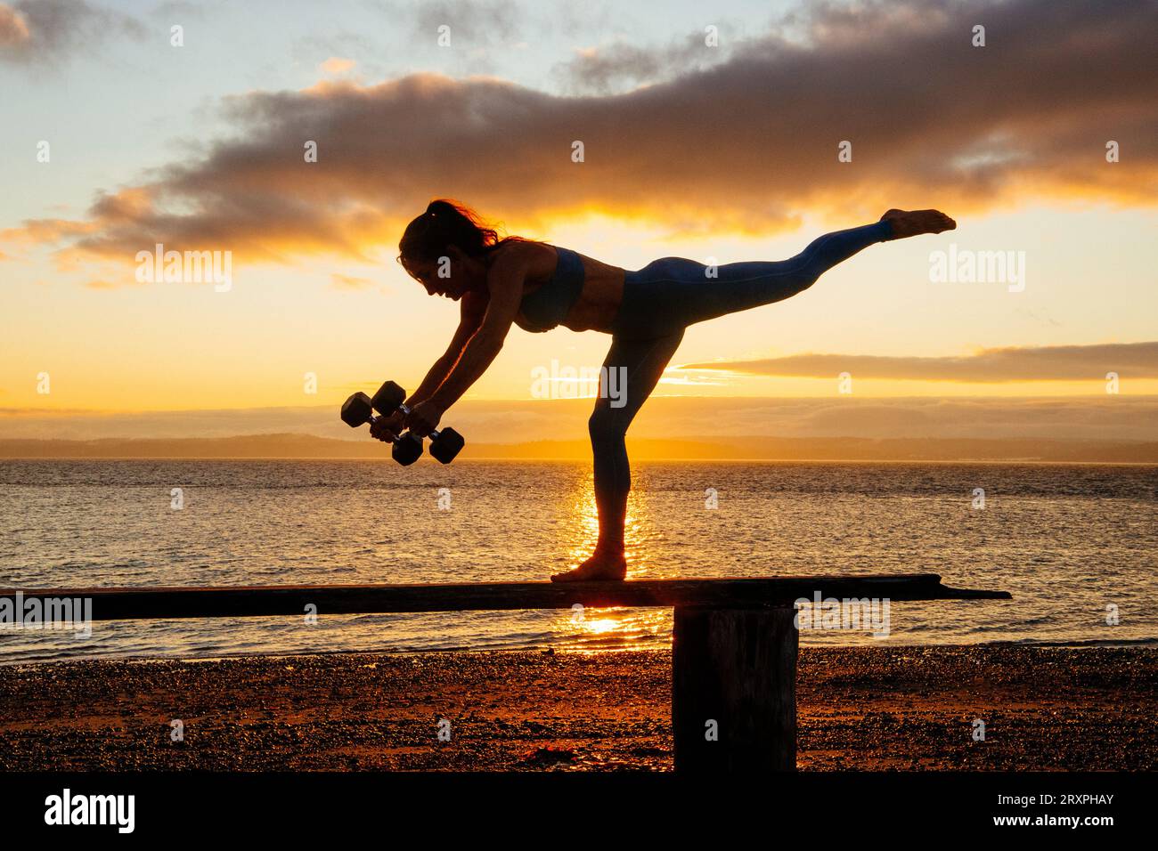 Silhouette de femme s'exerçant avec des haltères sur le dessus du banc côtier au crépuscule nuageux Banque D'Images