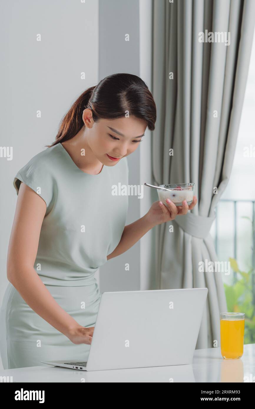 Femme satisfaite mangeant le petit déjeuner en utilisant un ordinateur portable sur un bureau à la maison Banque D'Images