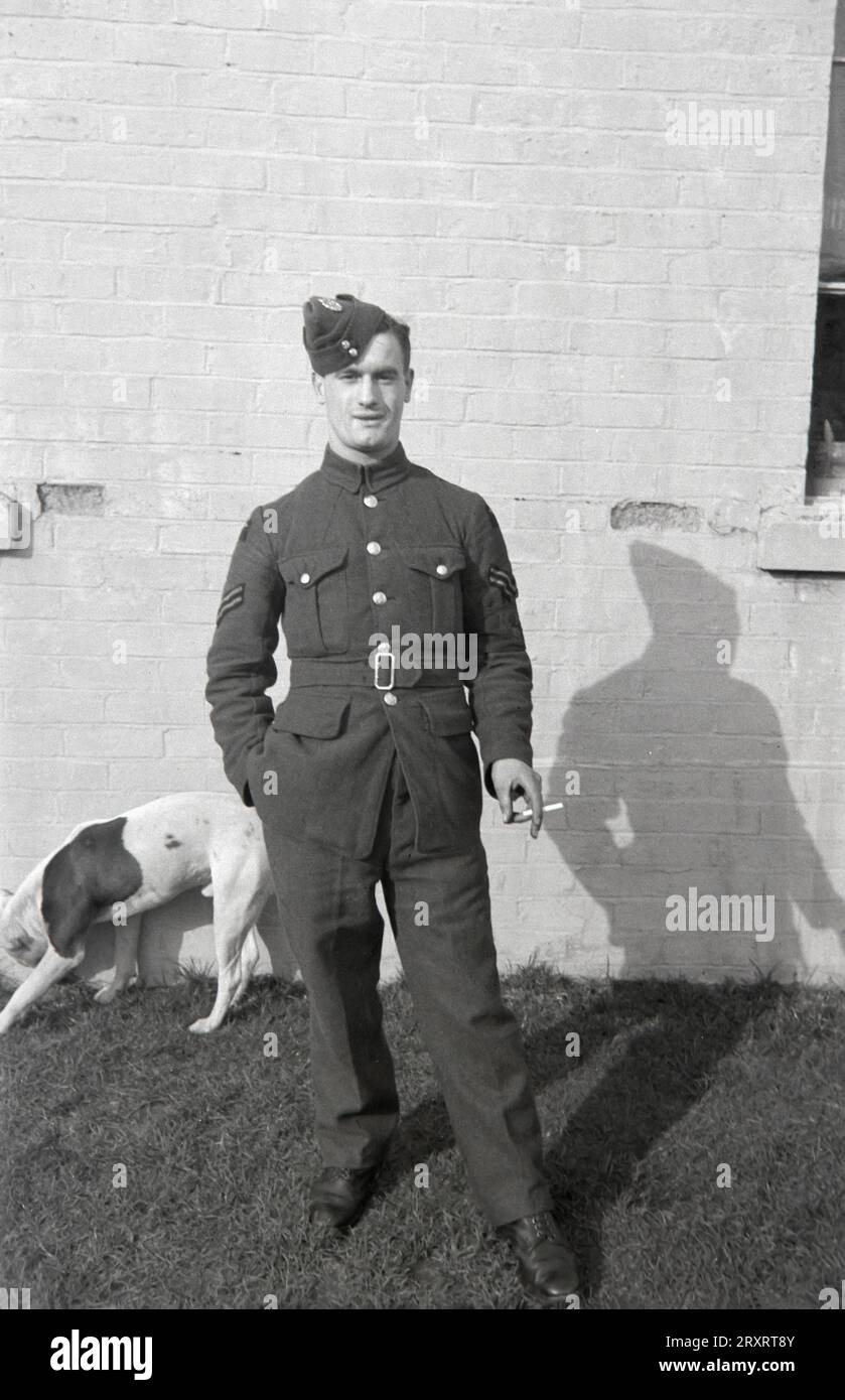 Une photographie vintage en noir et blanc des années 1930 montrant un aviateur britannique vêtu d'un uniforme d'époque, debout devant sa maison, fumant une cigarette. Banque D'Images