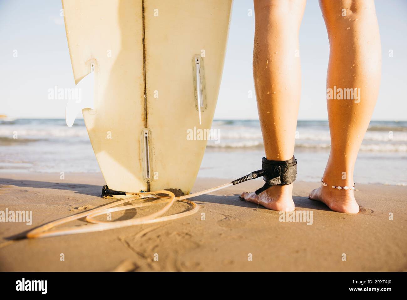 Gros plan des pieds d'une femme avec une planche de surf au bord de l'eau. Femme surfeuse attendant d'entrer dans l'eau pour profiter d'une journée de repos à la plage. Banque D'Images