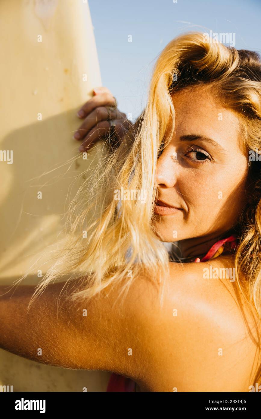 Portrait d'une surfeuse debout sur la plage en maillot de bain, avec sa planche de surf à ses côtés. Femme heureuse faisant des activités de plage pendant les vacances d'été. Banque D'Images