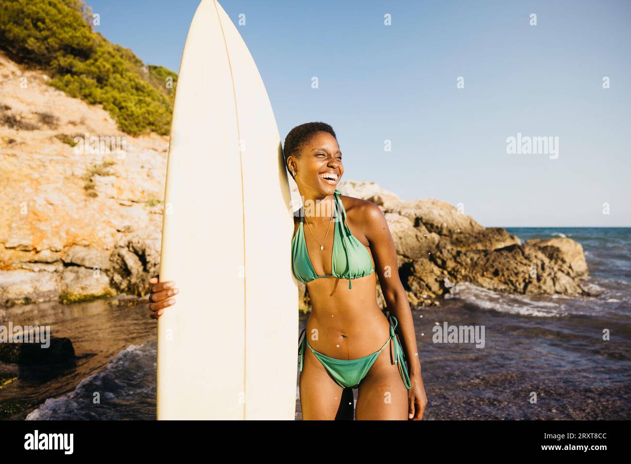 Portrait d'une surfeuse debout sur la plage en maillot de bain, avec sa planche de surf à ses côtés. Femme heureuse faisant des activités de plage pendant les vacances d'été. Banque D'Images