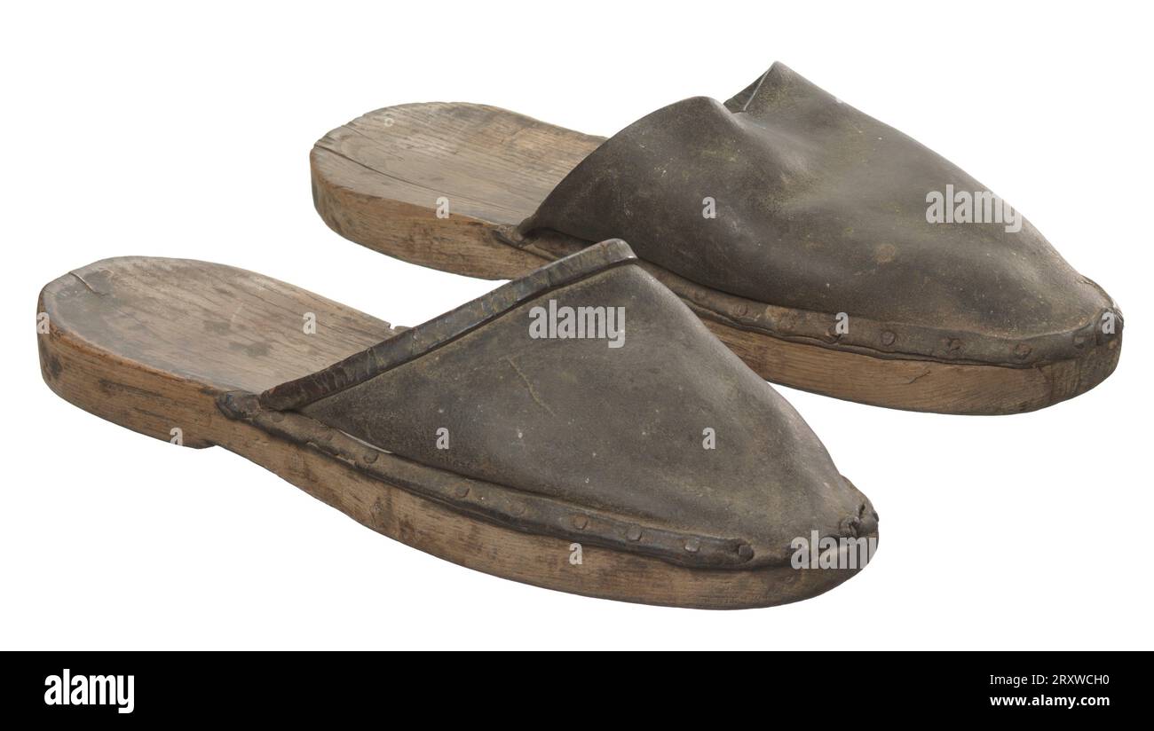 Une paire de chaussures mules ou pantoufles avec semelles en bois et dessus en cuir. Les semelles sont façonnées pour les pieds gauche (a) et droit (b). Les chaussures ont des orteils arrondis et des talons sculptés, légèrement relevés. Les chaussures ont seulement une empeigne, de sorte que le cuir ne pourrait pas entourer complètement le pied de l'utilisateur. Chaque empeigne est fabriquée à partir d'une seule pièce de cuir qui se double au point où l'empeigne se fixe à la semelle, ainsi qu'à l'ouverture du pied, pour plus de force. Les vamps sont accrochés aux semelles en bois avec des chevilles en métal. Les chaussures se glissent sur le pied du porteur et n'ont aucune méthode de fermeture. Banque D'Images