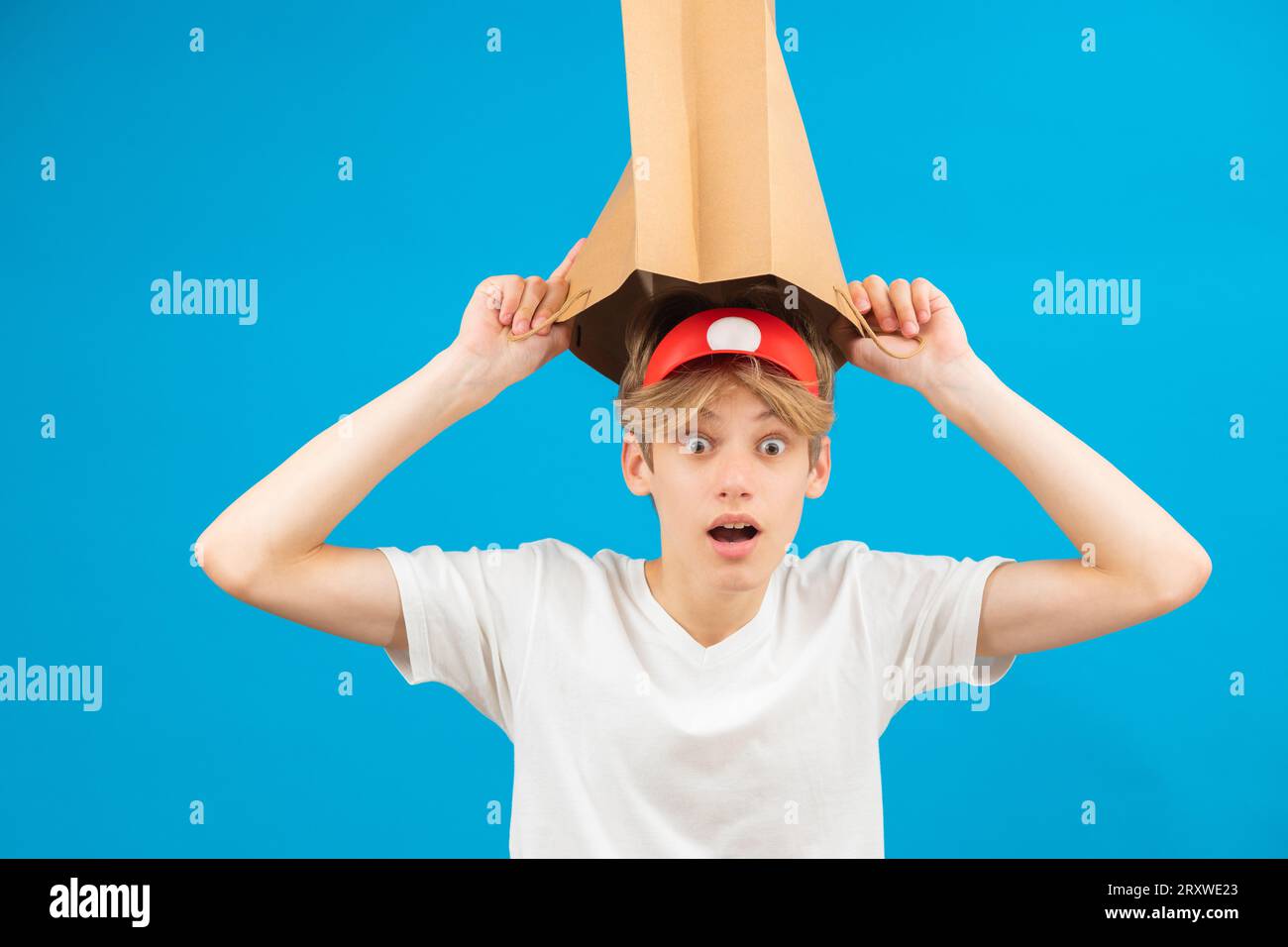 Garçon adolescent vendeur choqué avec sac en papier au-dessus de la tête. Adolescent tenant un sac en papier au-dessus de la tête posant en studio. Banque D'Images