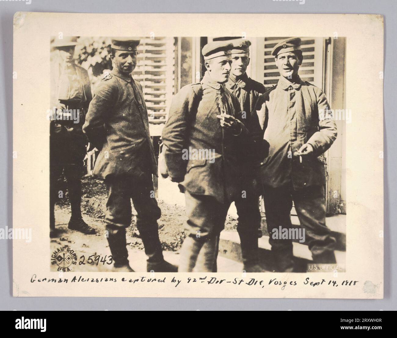 Cette photographie représente quatre soldats capturés debout devant un bâtiment avec des volets. À l'extrême gauche de la photo, un soldat portant un casque et une veste ouverte se tient à une courte distance des soldats capturés. Les soldats portent des bérets et des vestes fermées. Un des soldats à gauche regarde directement la caméra. Deux soldats tiennent des cigarettes dans leurs mains et le soldat debout derrière eux regarde la caméra. Le logo signal corps USA se trouve dans le coin inférieur gauche de la photo. Le numéro [25943] est écrit à côté du logo. Le mot Banque D'Images