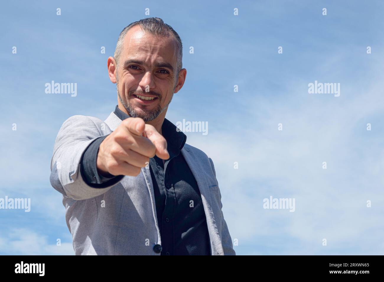 Homme d'affaires caucasien français avec barbe et vêtements d'affaires, debout à l'extérieur, heureux souriant pointant du doigt et regardant la caméra, backgro du ciel Banque D'Images