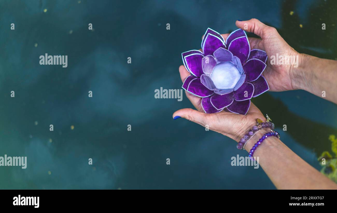 Vue d'angle de dessus des mains tenant un lotus en verre violet au-dessus de l'eau. Concept de yoga et de méditation. Espace de copie Banque D'Images