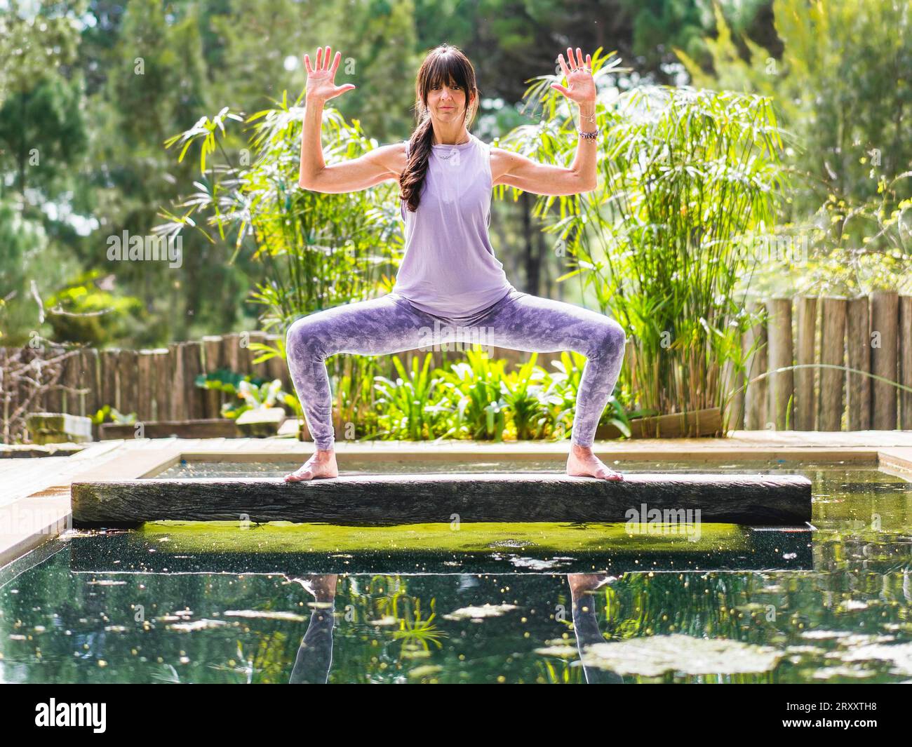 Une femme pratiquant le yoga pose Déesse avec Cactus Arms sur un tronc d'arbre au-dessus d'une piscine Banque D'Images