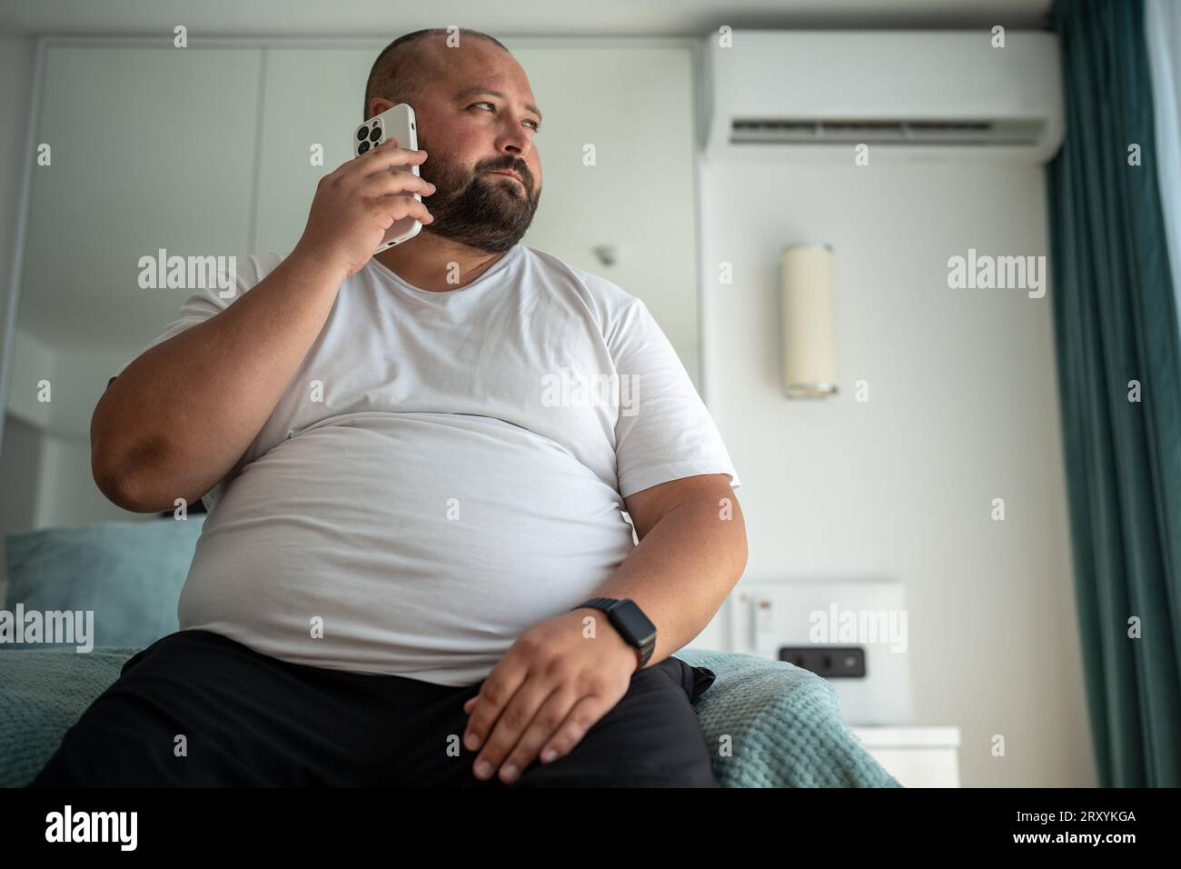 Gros gars appelle au téléphone, assis sur le lit, regardant par la fenêtre, s'inscrit à la salle de gym pour mettre la silhouette en ordre Banque D'Images