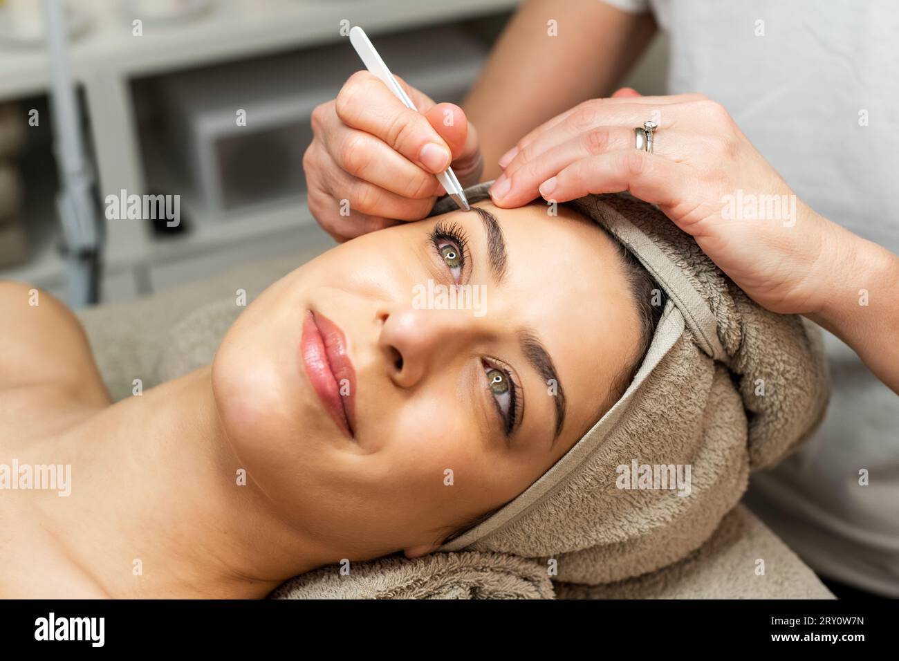 Grand angle de contenu dame avec serviette sur la tête couchée sur le lit tandis que la culture anonyme cosméticienne épilant les sourcils dans le salon de beauté Banque D'Images