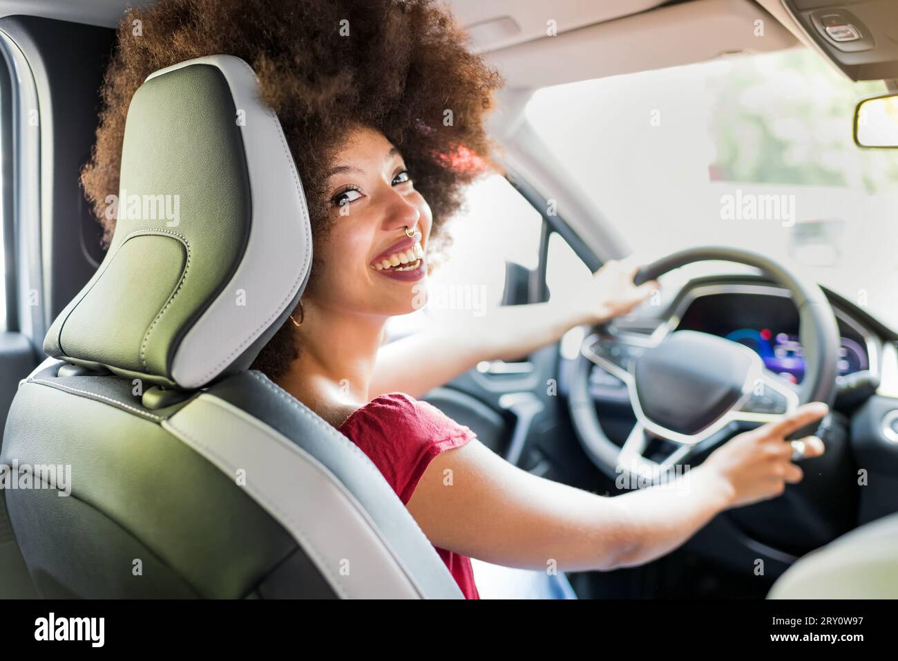 Heureuse femme afro-américaine avec coiffure afro conduisant l'automobile moderne tout en regardant loin avec sourire toothy Banque D'Images