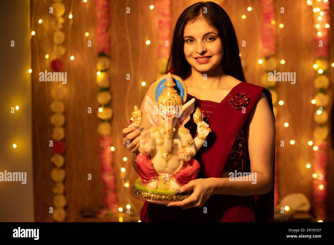 Heureuse jeune belle femme indienne en robe traditionnelle tenant Ganesh idole ou Ganesh Murti avec des lumières festives et décoration florale dans le backgroun Banque D'Images