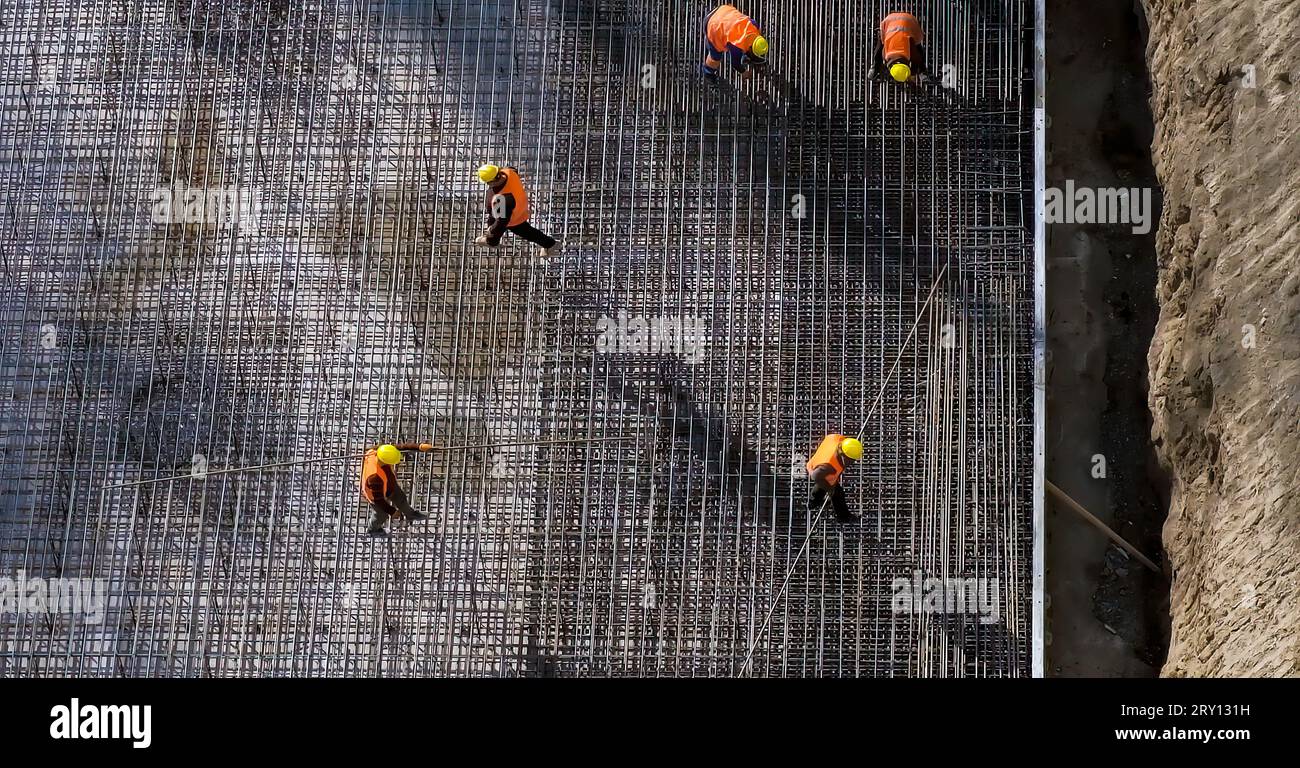 Chantier de construction. Ouvriers en uniforme orange portant des barres d'acier. Plancher de barres d'armature, constructeurs travaillant autour. Vue de dessus, lumière du jour. Photo de haute qualité Banque D'Images