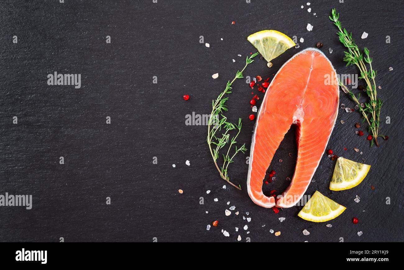 Steak de poisson de saumon cru frais avec des ingrédients de cuisine, des herbes et du citron sur fond noir, vue de dessus. Steak de saumon avec thym et citron sur table noire. Banque D'Images