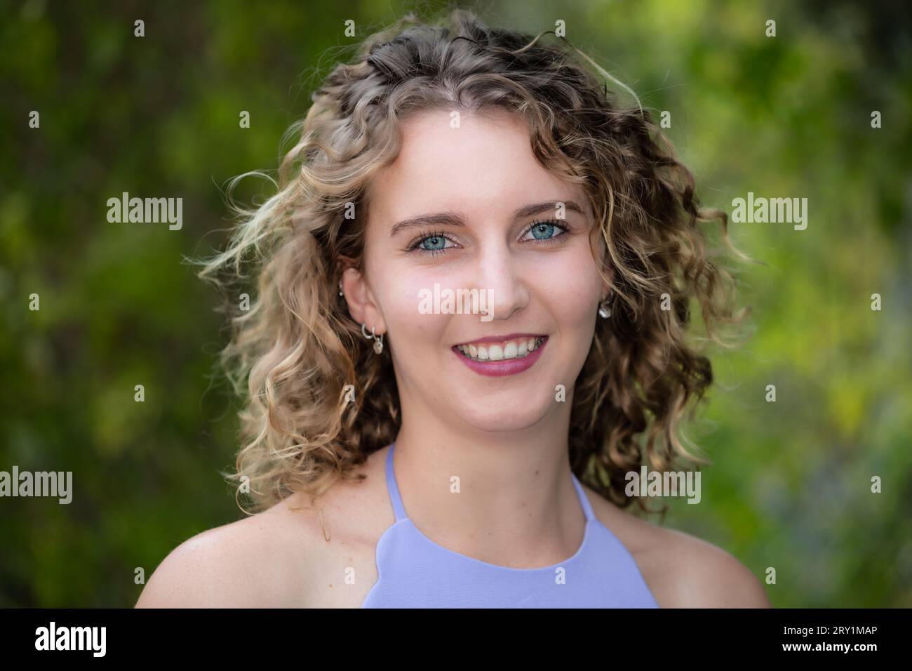 Portrait de la tête et des épaules d'une jeune femme caucasienne souriante aux cheveux blonds bouclés et aux yeux bleus, à l'extérieur en été Banque D'Images