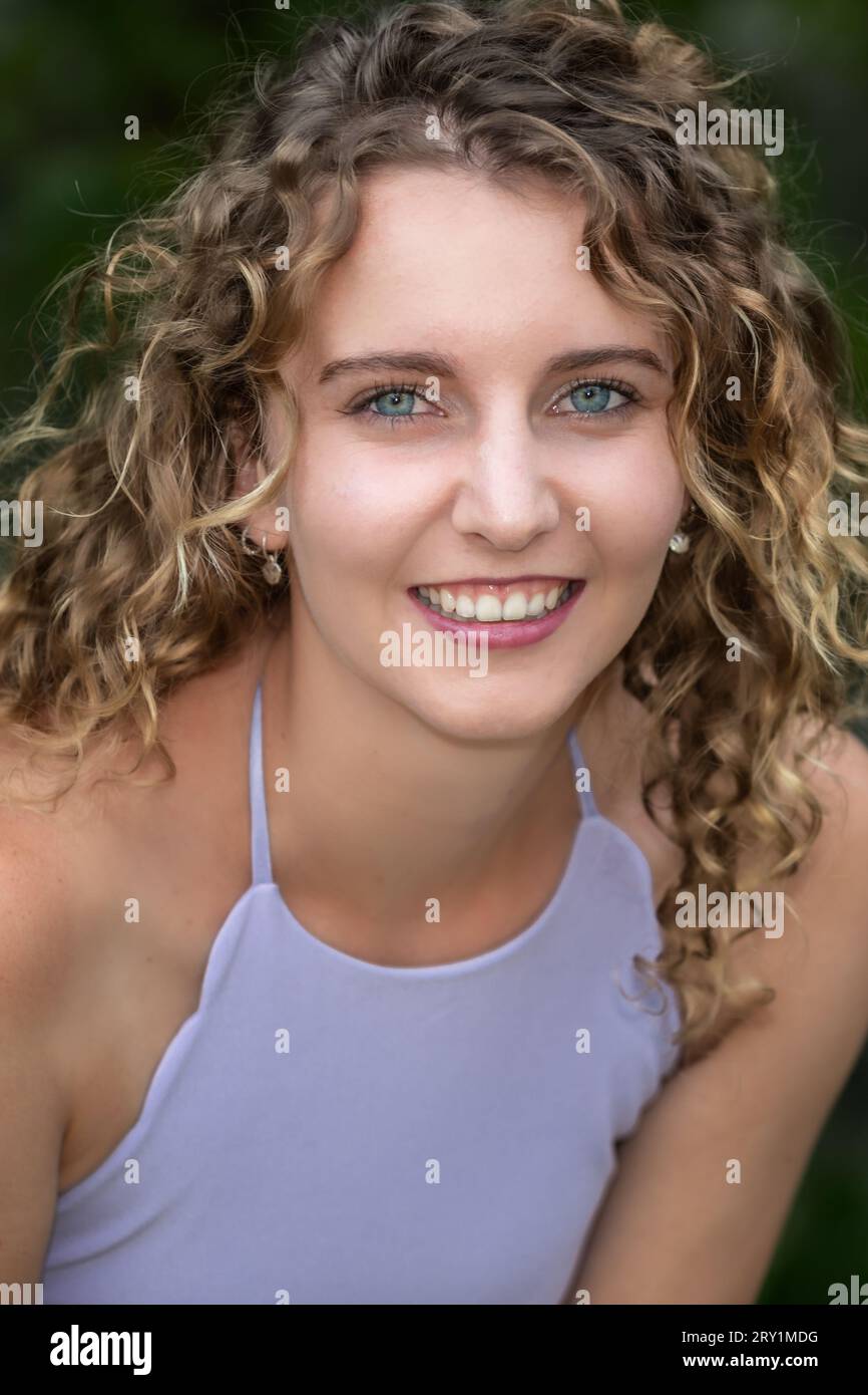 Portrait de la tête et des épaules d'une jeune femme caucasienne souriante aux cheveux blonds bouclés et aux yeux bleus, à l'extérieur en été Banque D'Images