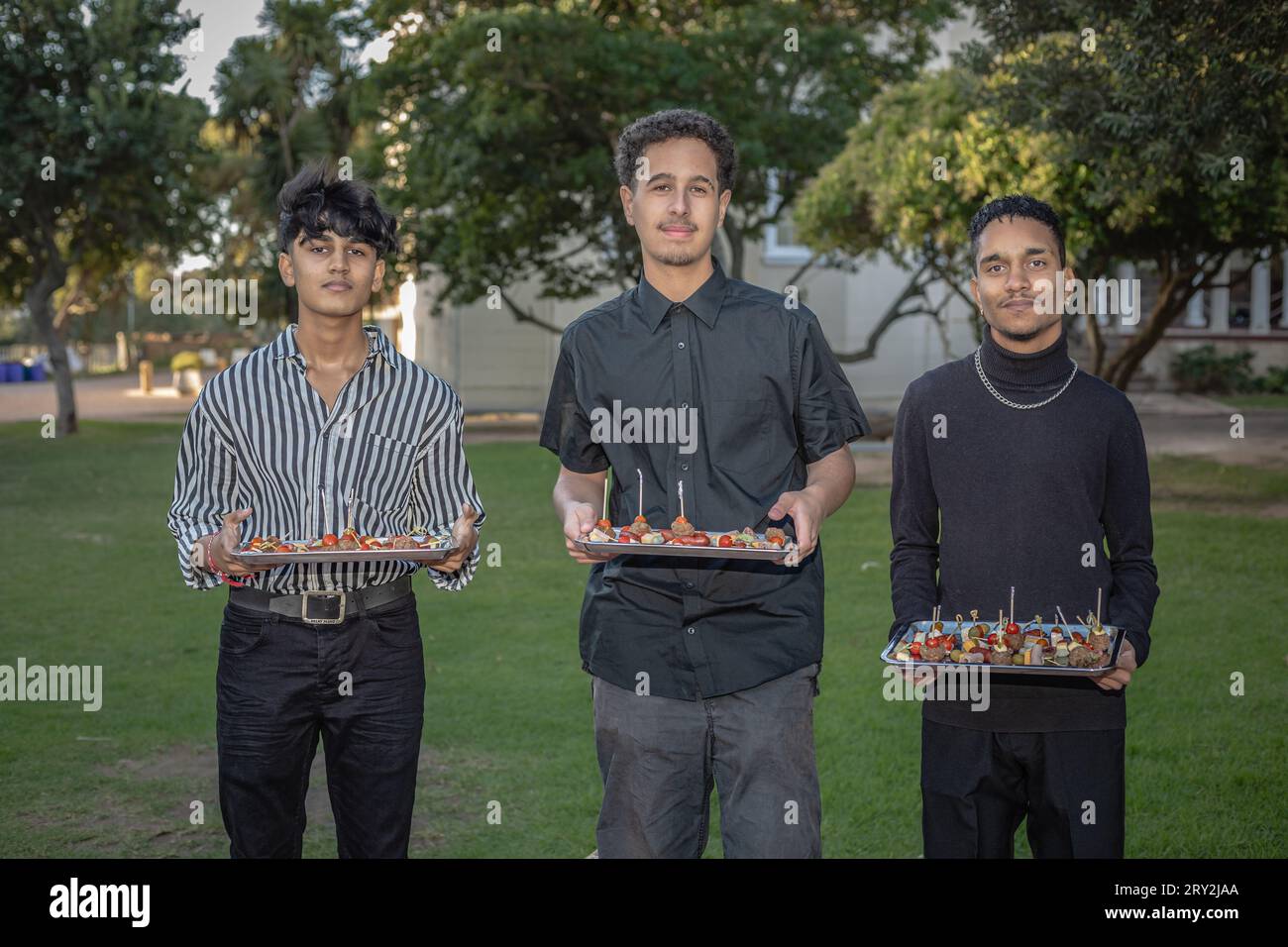 Au milieu de l'ambiance extérieure, trois jeunes serveurs de race mixte équilibrent habilement des plateaux chargés de délicieux canapés, incarnant le professionnalisme Banque D'Images