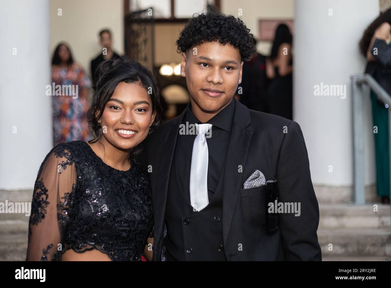 Dans un gros plan au bal de promo, un couple métis vêtu de vêtements habillés noirs élégants partage des sourires authentiques, dégageant élégance et bonheur Banque D'Images