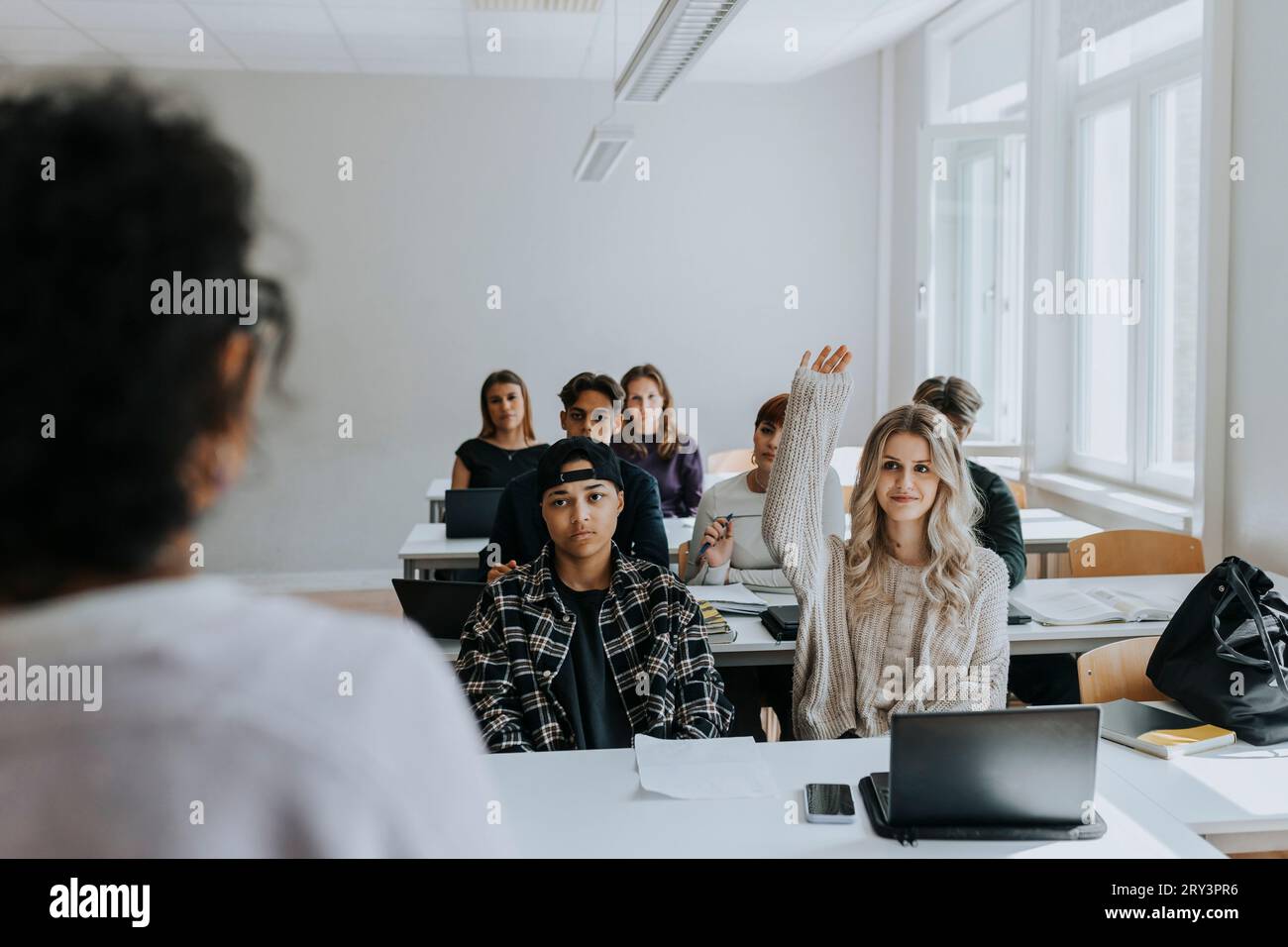 Jeune étudiante avec la main levée assise par des amis regardant l'enseignant dans la salle de classe Banque D'Images