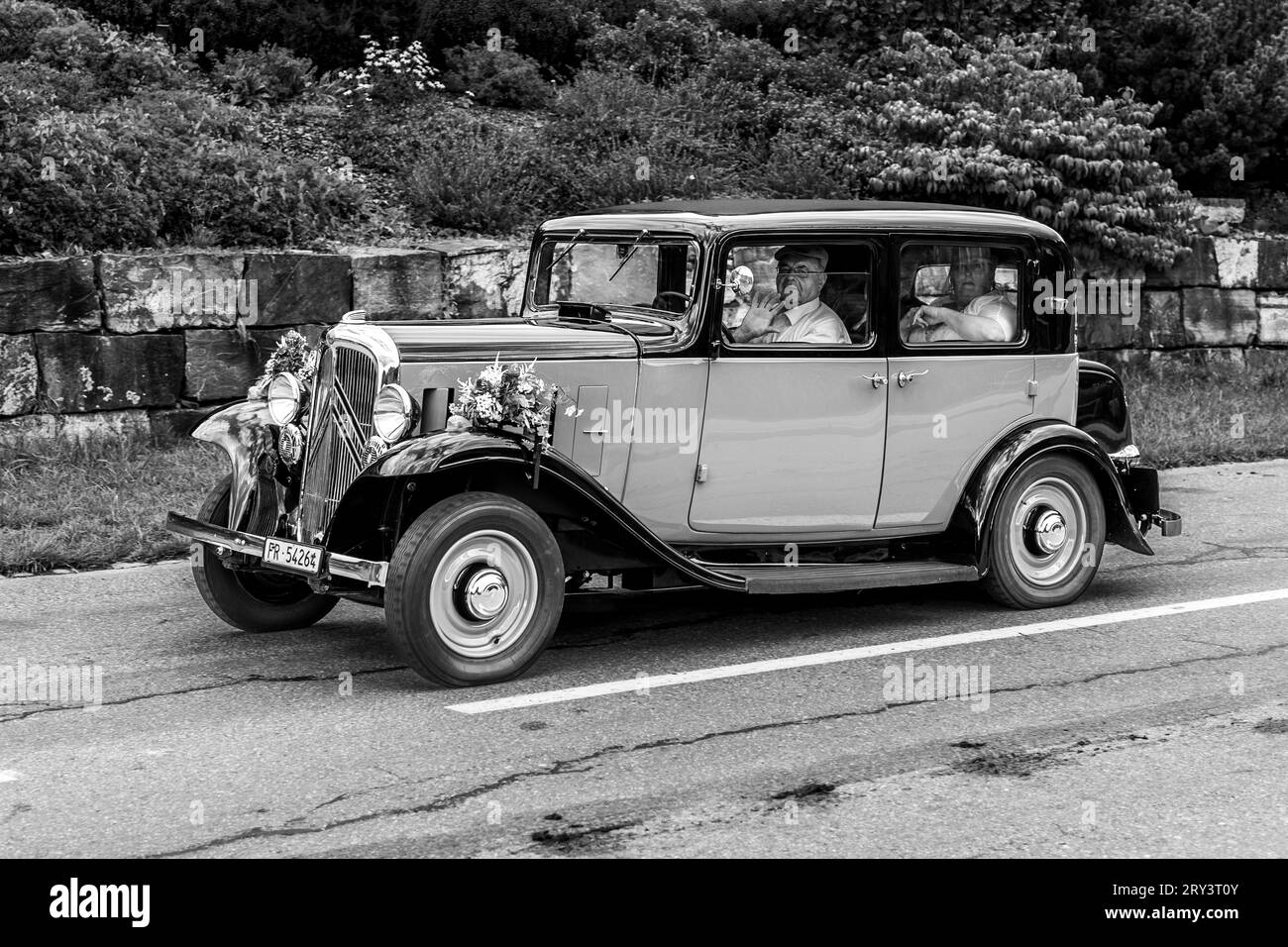 Une voiture vintage des années 1930, décorée festivement avec des fleurs blanches, transporte un couple marié devant un mur en pierre naturelle à Jaun, canton de Fribourg, Suisse. Un chauffeur portant un chapeau est assis au volant, la mariée avec un voile blanc est visible sur la banquette arrière. Banque D'Images