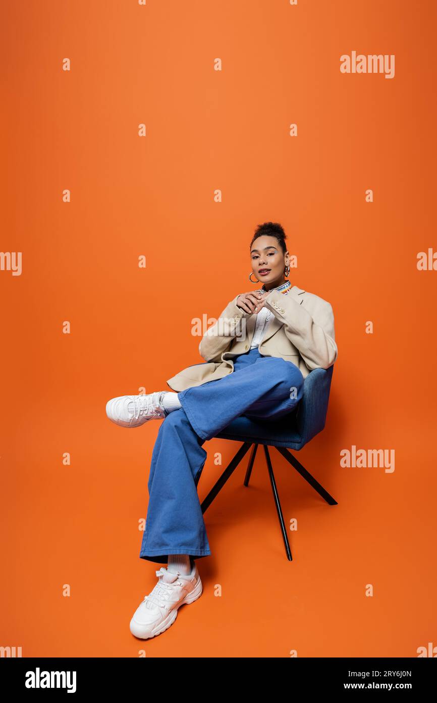 femme afro-américaine élégante en tenue à la mode assise sur une chaise bleue avec les mains plus près du visage Banque D'Images