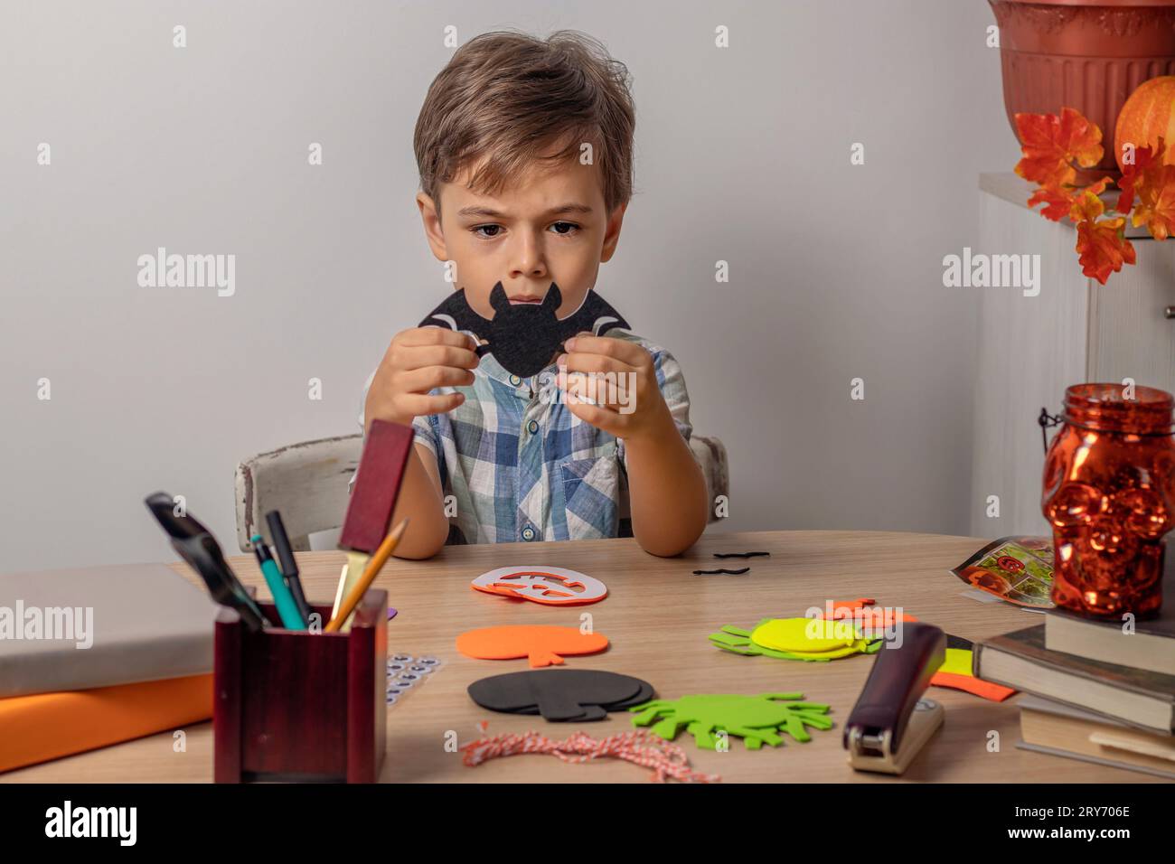 mignon garçon est assis à la table et tient une chauve-souris noire fabriquée dans ses mains. Sur la table sont de la corde, des citrouilles sculptées, des araignées et des chauves-souris. Préparation pour Halloween. Artisanat d'automne ou concept DIY Banque D'Images